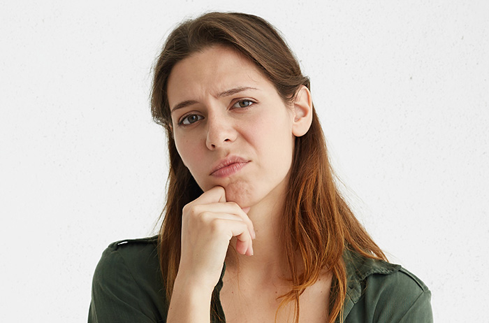 Vorteile für Sie durch den S-Pap könnten Nachteile für Ihren Frauenarzt bedeuten. Es gibt verschiedene, mögliche Gründe, warum Ihr Frauenarzt den S-Pap ablehnen könnte. Ihr Menschenrecht auf Selbstbestimmung wird dabei nicht beachtet. Junge Frau mit braunen langen Haaren in grünem T-Shirt vor grauem Hintergrund hat ihr Kinn auf der rechten Hand abgestützt und schaut besonders skeptisch und mismutig in die Kamera.