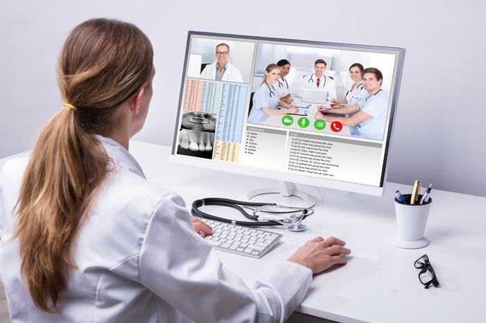 Auch beim Informationsaustausch streben wir seit Jahren einen hohen Standard an. Die viefältigen technischen Möglichkeiten helfen bei der Beratung und schaffen Sicherheit bei Auffälligkeiten und Unklarheiten. Bei auffälligen Befunden ermöglichen unsere Fallkonferenzen die notwendige Klarheit, um vernünftige Therapieentscheidungen zu treffen. Ärztin mit langen blonden Haaren als  Pferdeschwanz im Kittel sitz vor weißem Schreibtisch mit Bildschirm, auf dem Ärzte als Team-Konferenz  zu sehen sind.
