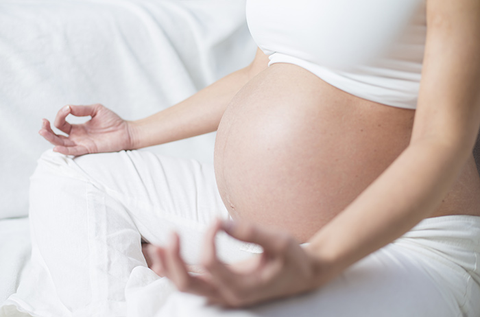 Kinderwunsch unterstützen und Frühgeburtlichkeit vermeiden Seitliches Abbild einer meditierenden schwangeren Frau von Brust bis Fuß, bauchfrei mit weißem Oberteil und weißer Hose, die Hände auf den Beinen liegend und Daumen und Zeigefinger meditativ aneinander haltend.