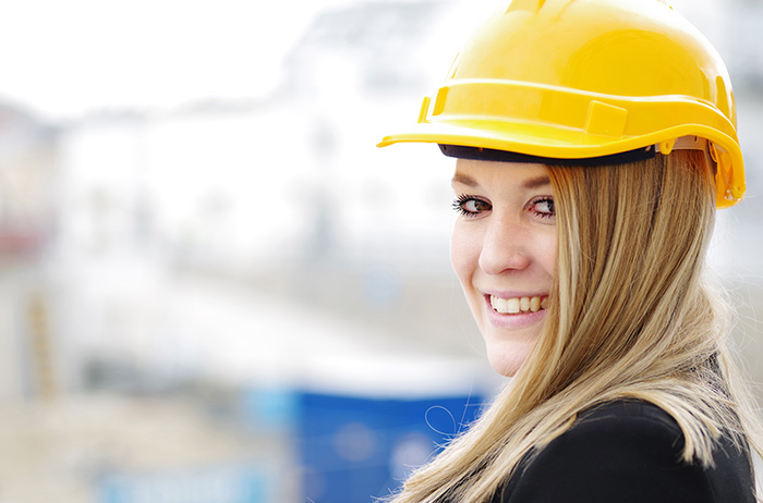 Kolleginnen in Unternehmen und Organisationen schützen Junge, blonde Frau mit einem gelben Baustellen-Helm auf dem Kopf lächelt seitlich in die Kamera.