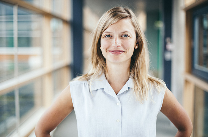 Informationen zu Gebärmutterhalskrebs sind oft fehlerhaft. Statt blindem Vertrauen sollte das Arzt-Patienten-Verhältnis durch Zusammenarbeit auf Augenhöhe stattfinden. Sie können Eigenverantwortung übernehmen und wir helfen Ihnen dabei. Junge blonde Frau mit weißer kurzärmliger Bluse steht in einem Gebäude, die Hände an den Hüften positioniert, und lächelt selbstbewusst in die Kamera.