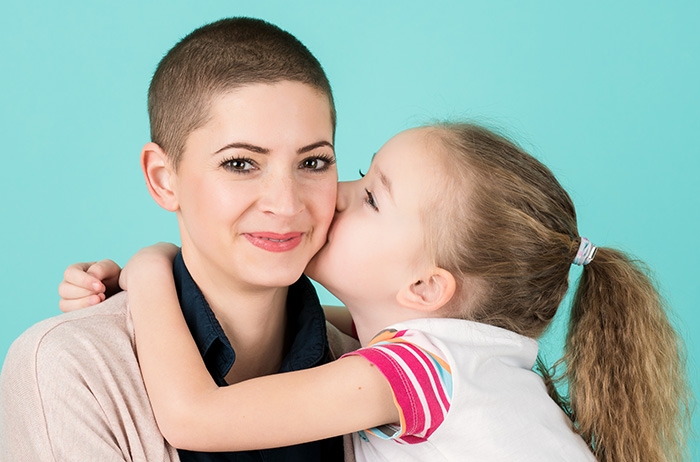 Sichere Überwachung nach allen anderen Krebsdiagnosen Kleines Mädchen mit Zopf küsst ihre lächelnde Mutter mit kurzen Haaren, die eine Krebspatientin ist/war, auf die Wange.