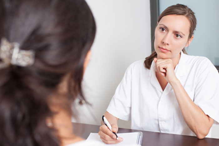 Die Sicherheit, Vorstufen von Gebärmutterhalskrebs bei der Vorsorge zu erkennen, ist abhängig von Abstrichmethode. Neben dem konventionellen Pap-Test und der Dünnschichtzytologie gibt es jetzt auch den S-Pap mit höchster Sicherheit. Ärztin in weißem Kittel am Schreibtisch sitzend mit Kugelschreiber und Schreibblock berät konzentriert eine Patientin, die ihr gegenüber sitzt und von der nur nur braune Haare mit Haarspange im linken Bildrand vonhinten zu sehen sind.