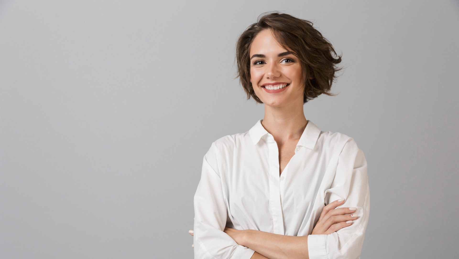 Wir tun alles um Ihnen zu helfen, gesund zu werden und gesund zu bleiben. Mit verlässlichen Informationen, Angeboten, Rat und Tat. Junge Frau mit kastanien-braunen Haaren und weisser Bluse steht vor grauem Hintergrund mit verschränkten Armen und lächelt optimistisch  als wolle sie sagen: Ich feue mich über Ihren Besuch!